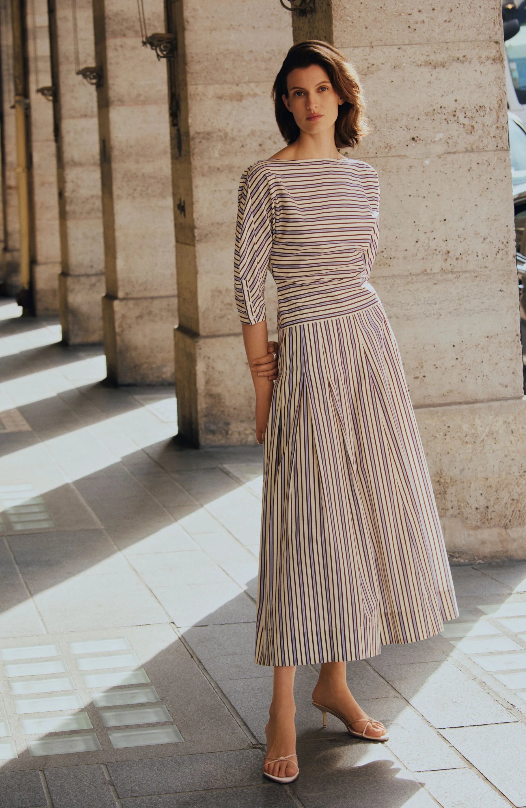 A woman stands on a sunlit stone walkway in chic workwear—a Lela Rose Striped Cotton Dolman Sleeve Top styled as a dress with open-toed heels—columns rising elegantly in the background.