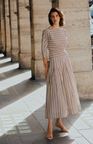 A woman stands on a sunlit stone walkway in chic workwear—a Lela Rose Striped Cotton Dolman Sleeve Top styled as a dress with open-toed heels—columns rising elegantly in the background.