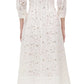 A person shown from the back wears Simkhai’s Jazz Floral Broderie Anglaise Dress—a white, long-sleeved, calf-length lace dress with a fitted waist—paired with white heeled sandals, exuding classic femininity through delicate floral details.