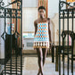 A woman wearing the Cara Cara Valencia Mini Dress stands barefoot in a stylish, well-lit room featuring striped curtains, black iron gates, and decorative accents.