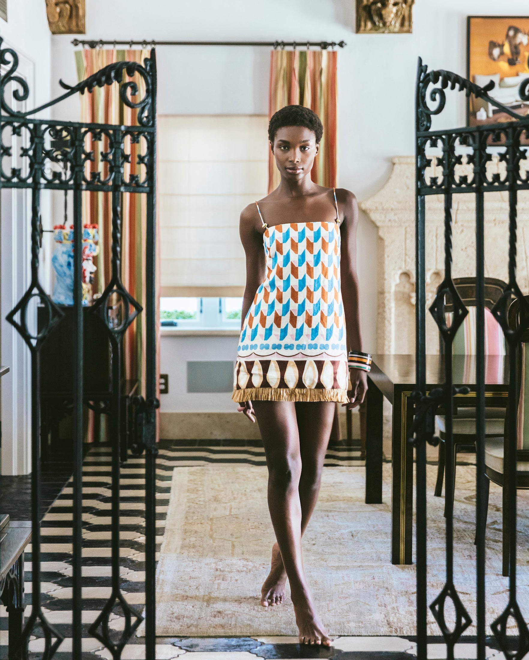 A woman wearing the Cara Cara Valencia Mini Dress stands barefoot in a stylish, well-lit room featuring striped curtains, black iron gates, and decorative accents.