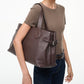 A person carries the Staud Piper Tote, a large brown leather work bag with buckle detailing, on their shoulder while wearing a brown short-sleeve top and dark jeans against a plain background.
