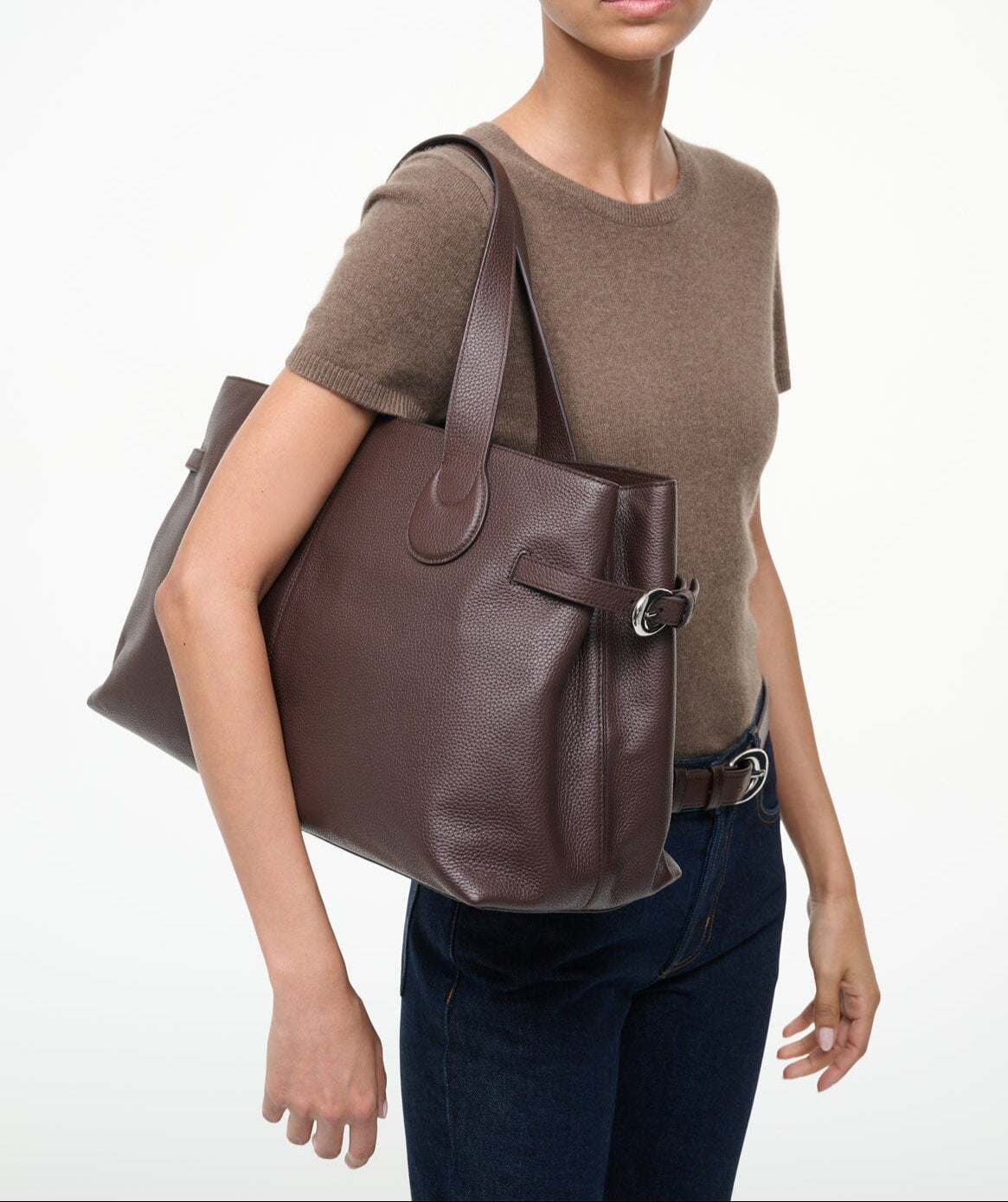 A person carries the Staud Piper Tote, a large brown leather work bag with buckle detailing, on their shoulder while wearing a brown short-sleeve top and dark jeans against a plain background.