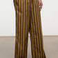 A person stands against a plain background wearing the Staud Mateo Cotton Poplin Pant—brown, wide-leg, and mid-rise with vertical yellow and gray stripes. The pants feature a comfortable drawstring waist, side pockets, and are crafted from breathable cotton poplin.