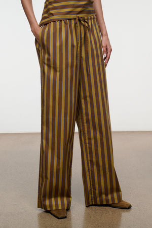 A person stands against a plain background wearing the Staud Mateo Cotton Poplin Pant—brown, wide-leg, and mid-rise with vertical yellow and gray stripes. The pants feature a comfortable drawstring waist, side pockets, and are crafted from breathable cotton poplin.