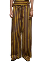 A person wearing the Staud Mateo Cotton Poplin Pant, featuring a mid-rise fit, drawstring waist, and brown and yellow vertical stripes in wide-leg cotton poplin, stands against a plain background.