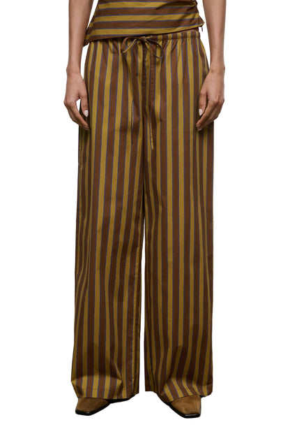 A person wearing the Staud Mateo Cotton Poplin Pant, featuring a mid-rise fit, drawstring waist, and brown and yellow vertical stripes in wide-leg cotton poplin, stands against a plain background.