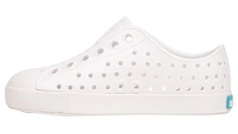 Native Shoes Kids’ Jefferson Shoes are white slip-on sneakers featuring round perforations, a white sole, and a small blue label on the heel. Shown in profile against a white background, these lightweight shoes are designed for comfort and everyday play.