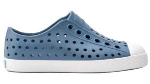 The Kids’ Jefferson Shoes by Native Shoes feature a blue slip-on design with circular ventilation holes, a white toe cap, and a white sole, shown here in side profile. Lightweight and water-friendly, they’re perfect for kids’ everyday adventures.