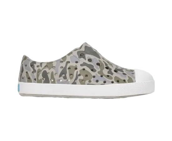 A side profile view of the Kids’ Jefferson Print Shoes by Native Shoes is displayed against a white background. This gray camouflage slip-on sneaker, crafted with Sugarlite™ technology, features a shock-absorbent white sole and toe cap, offering lightweight comfort and a modern design.