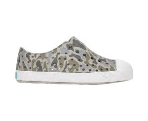 A side profile view of the Kids’ Jefferson Print Shoes by Native Shoes is displayed against a white background. This gray camouflage slip-on sneaker, crafted with Sugarlite™ technology, features a shock-absorbent white sole and toe cap, offering lightweight comfort and a modern design.