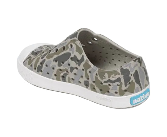 The Kids’ Jefferson Print Shoes by Native Shoes feature a green and gray camouflage pattern, a white sole made from shock-absorbent Sugarlite material for extra comfort, ventilation holes for breathability, and a blue “native” label on the heel.