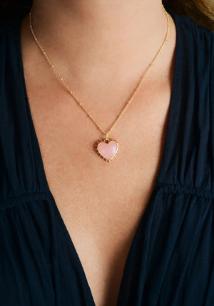 A person wears the Anabel Aram Heart with Stone Charm necklace, featuring a rose quartz heart pendant, paired with a deep v-neck dark blue top.
