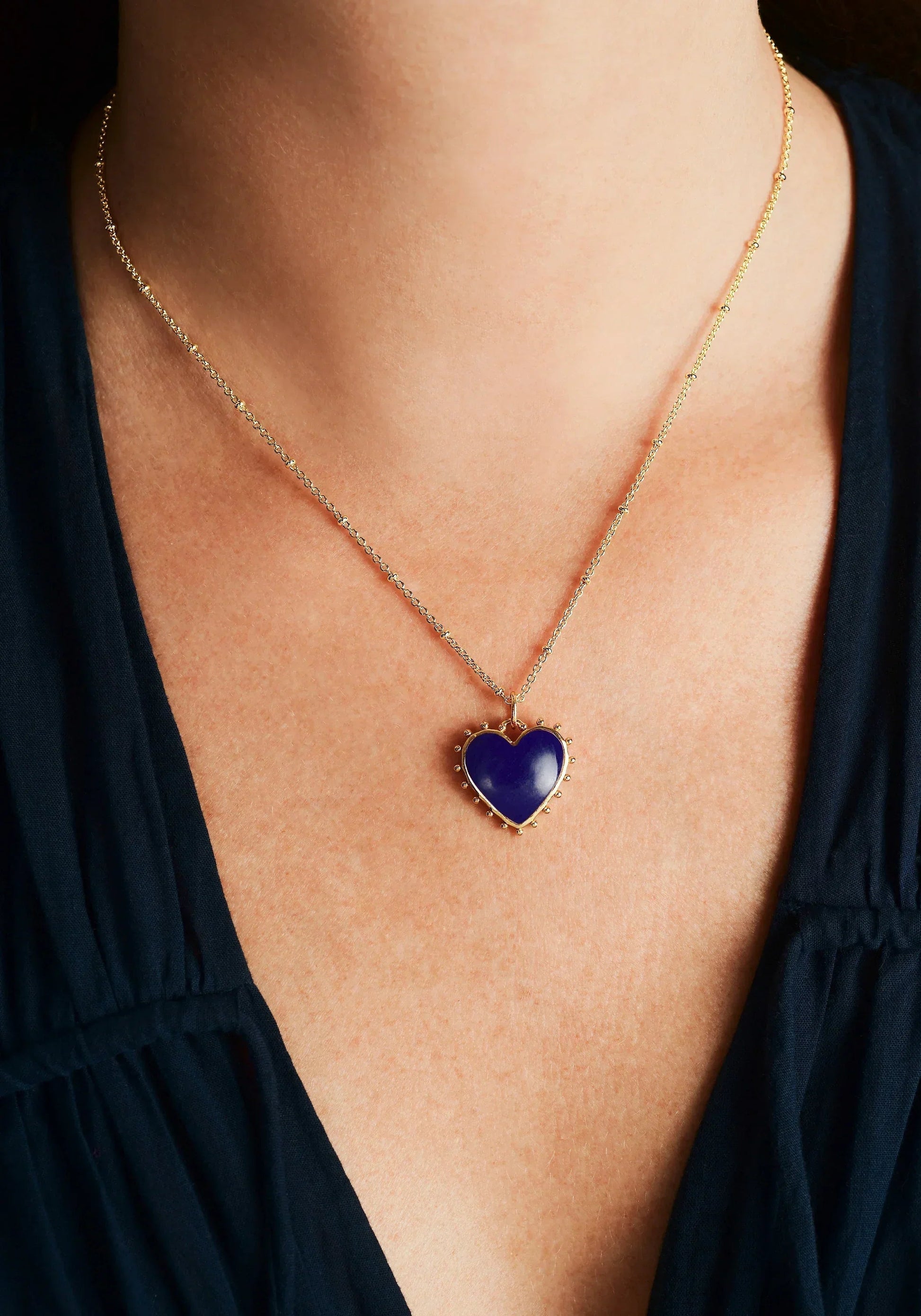 A person wears the Anabel Aram Heart with Stone Charm gold necklace featuring a blue heart-shaped pendant, styled against a dark V-neck top.