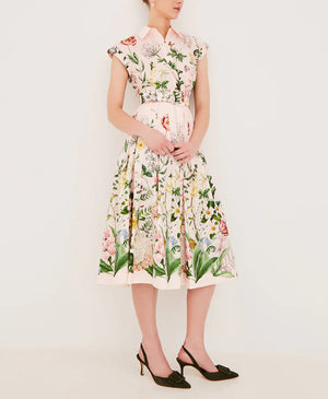 A woman stands against a plain wall in the Cara Cara Octavia Dress, a light pink floral fit-and-flare midi with a pleated skirt and collared neckline, highlighting her silhouette as she clasps her hands in front of her. She wears black heels.