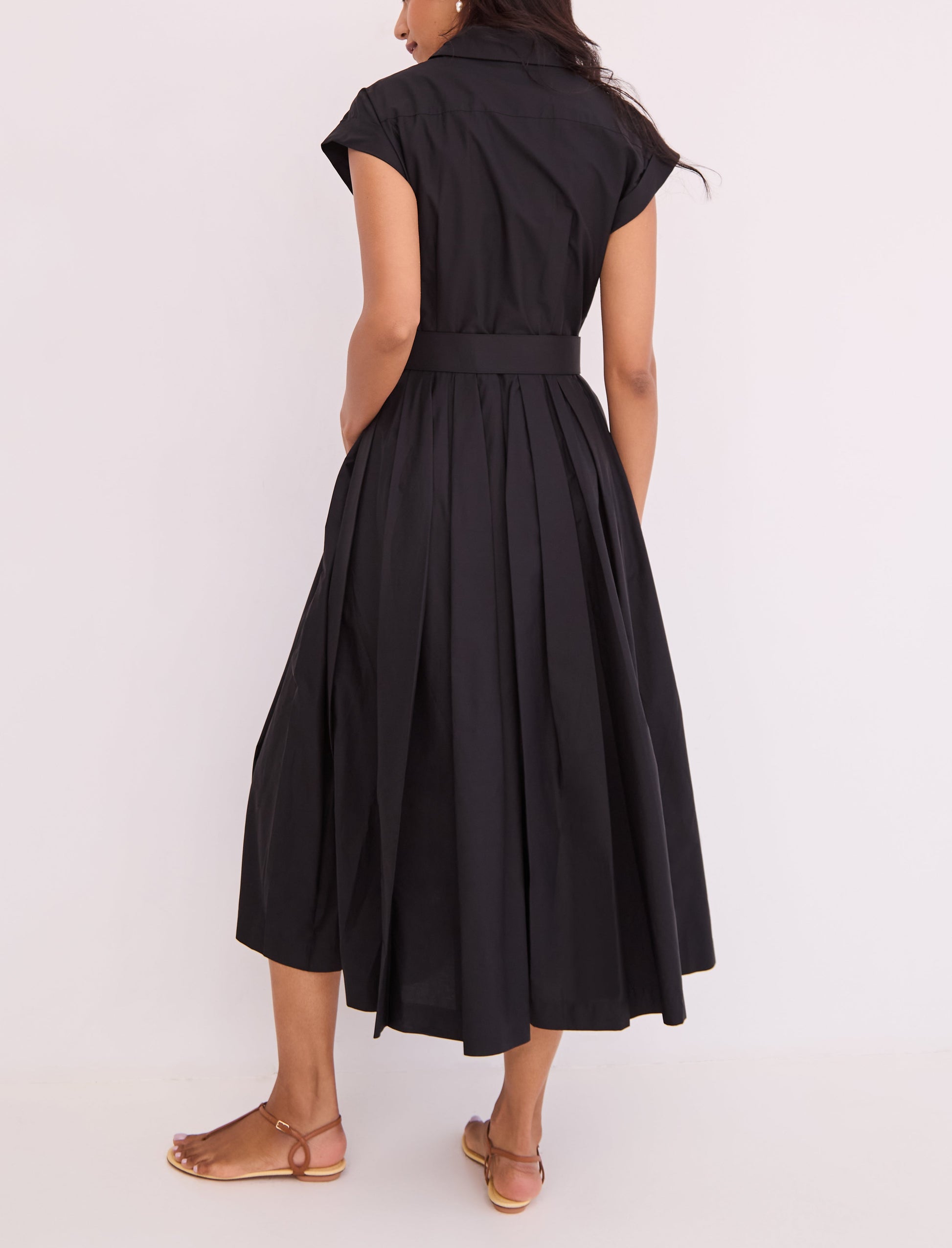 A person shown from the back wears the Octavia Dress by Cara Cara—a short-sleeve, collared, black belted shirtdress with a box pleat skirt—paired with flat sandals.