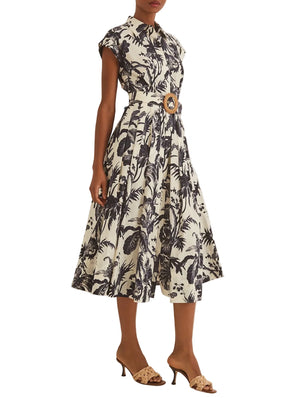 A person wears the Octavia Dress by Cara Cara, featuring a crisp belted silhouette with black palm tree print and short sleeves, paired with tan heeled sandals—a polished, flirtatious look for any occasion.