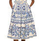 A woman wears the Cara Cara Octavia Dress, a crisp white sleeveless midi with blue floral and geometric prints, paired with flat brown sandals.