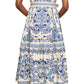 A woman faces away, wearing the Cara Cara Octavia Dress—a cream midi dress with blue florals, belted waist, cap sleeves, and flat sandals.