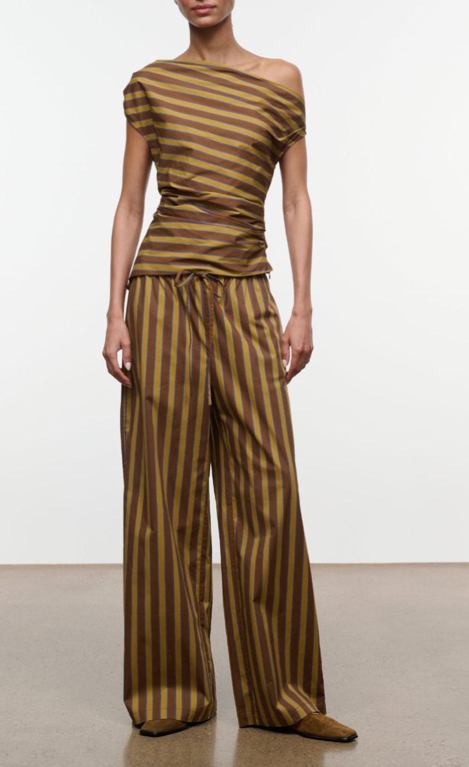 A person wearing the Staud Mateo Cotton Poplin Pant, a wide-leg, mid-rise style with a drawstring waist in a brown and yellow vertical stripe pattern, paired with a matching off-shoulder cotton poplin top, stands on a smooth indoor floor against a plain background.