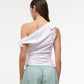 A person faces away, wearing the Staud Bellaria Top—a white one-shoulder evening top with an asymmetrical neckline and tied sleeve—paired with light green shorts against a plain background.