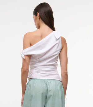A person faces away, wearing the Staud Bellaria Top—a white one-shoulder evening top with an asymmetrical neckline and tied sleeve—paired with light green shorts against a plain background.