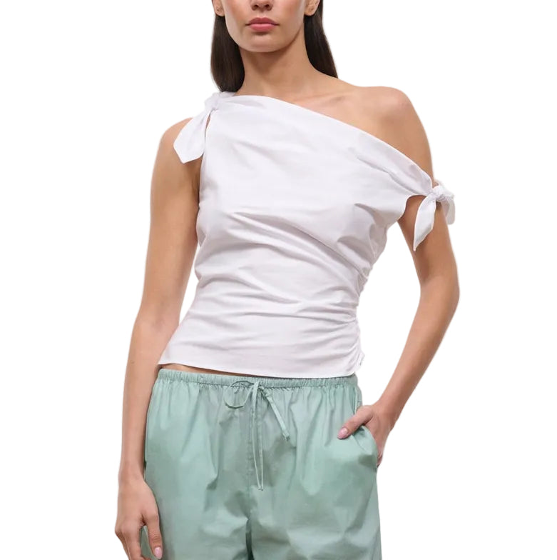 A woman wears the Staud Bellaria Top, a white one-shoulder evening top with an asymmetrical neckline and tie sleeve details, paired with light green drawstring pants.