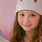 A young girl with long brown hair smiles at the camera, wearing the Great Pretenders Ombre Sequins Crown and holding a silver balloon against a pale pink background.