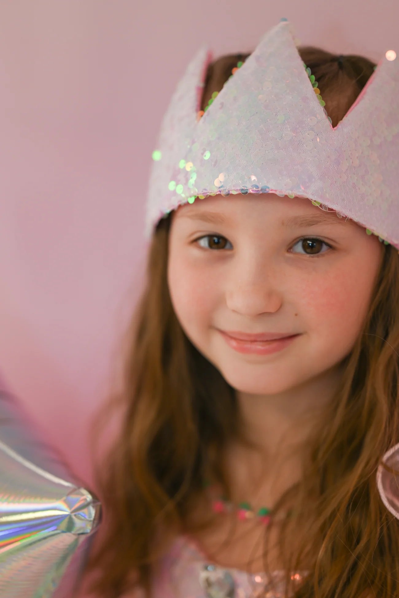 A young girl with long brown hair smiles at the camera, wearing the Great Pretenders Ombre Sequins Crown and holding a silver balloon against a pale pink background.