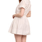 A woman models the Cara Cara Asbury Mini Dress—a short-sleeved, collared, light-toned shirtdress with a functional tie detail—standing angled to the side against a plain white background.