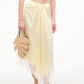A person in a white crop top and pale yellow lace-trim skirt holds the Staud Palm Raffia Bag, a leaf-shaped woven style with leather handles, and wears white sandals.