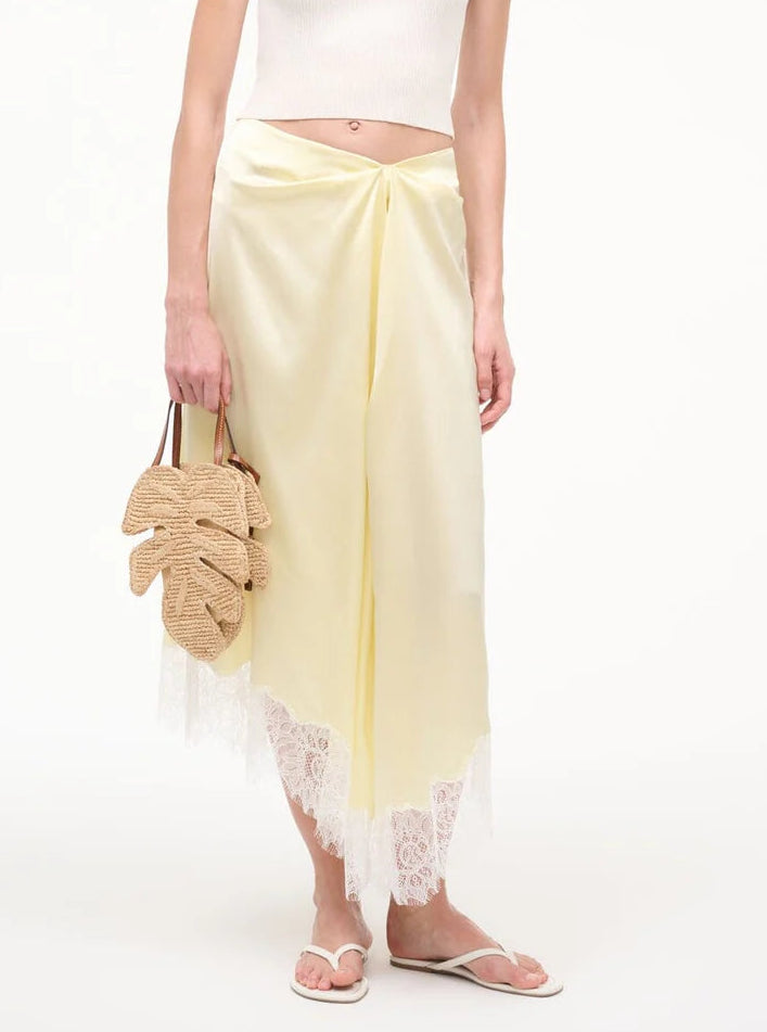 A person in a white crop top and pale yellow lace-trim skirt holds the Staud Palm Raffia Bag, a leaf-shaped woven style with leather handles, and wears white sandals.