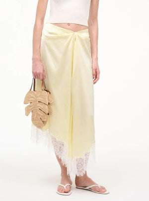 A person in a white crop top and pale yellow lace-trim skirt holds the Staud Palm Raffia Bag, a leaf-shaped woven style with leather handles, and wears white sandals.