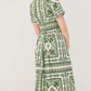 A woman is seen from the back wearing the Cara Cara Malena Dress, a green and white patterned short-sleeve dress with a belted, fitted waist, set against a plain background.