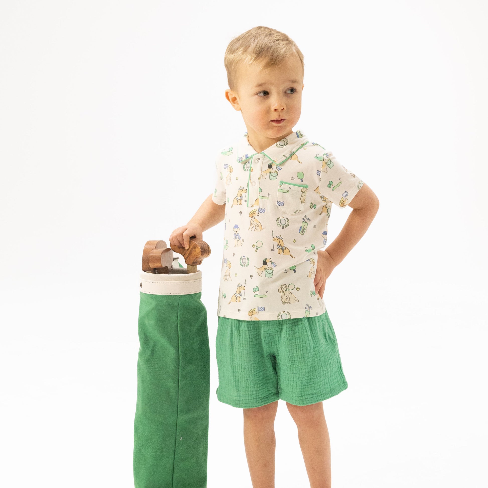 A young boy in the Angel Dear Baby Boys’ and Boys’ Polo with Casual Short stands next to a green golf bag, holding a wooden golf club against a plain white background. His organic cotton muslin shorts from Angel Dear offer both comfort and style for this sporty ensemble.