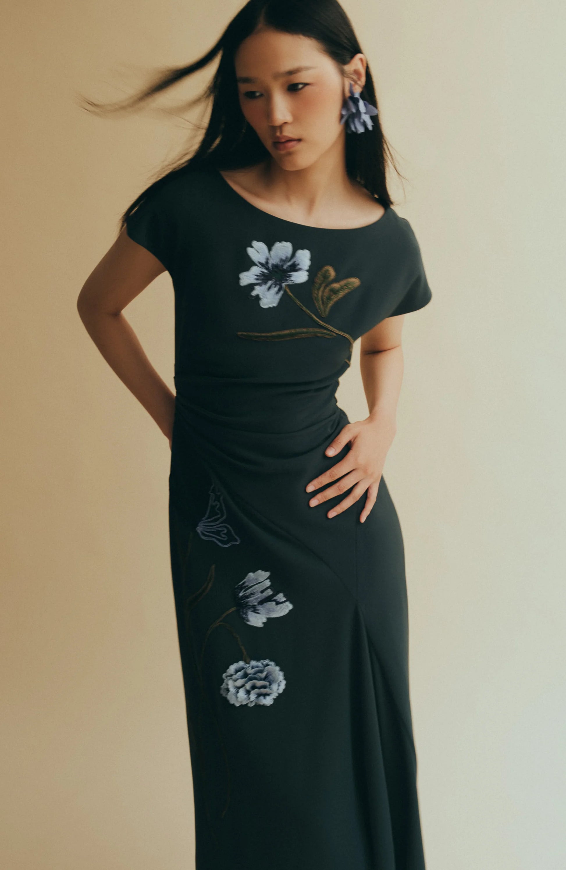 A woman poses against a beige background in the Lela Rose Floral Fluid Crepe Cascade Dress, featuring floral cord embroidery, with her hand on her hip and hair gently blowing.