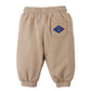 The Gingersnaps Baby Boys’ Fiulio Joggers are tan jogger pants featuring a blue patch on the back pocket, cozy pintuck details, and an elastic waistband and cuffs, displayed on a white background.