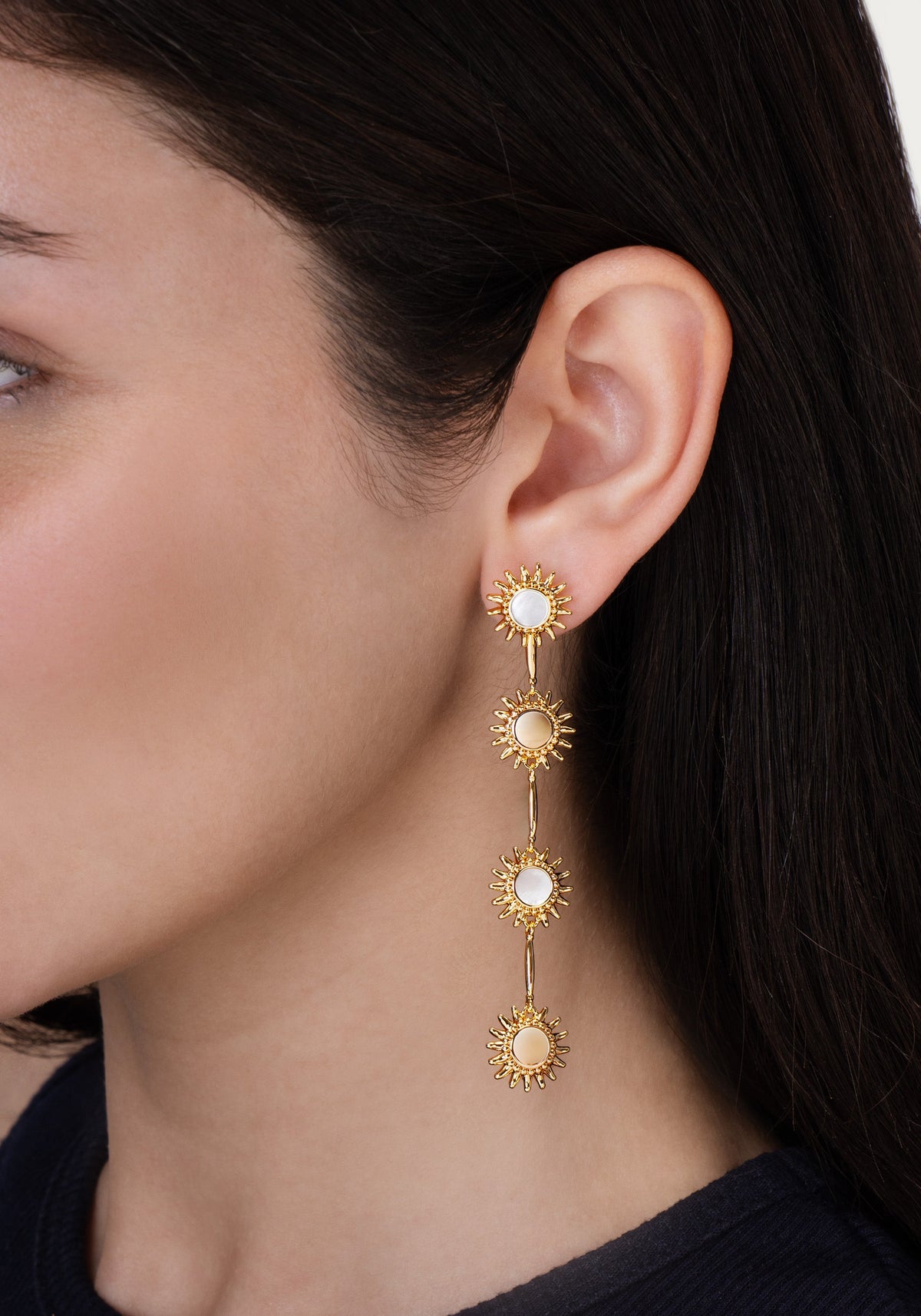 Anabel Aram Milos Soleil drop earrings in 18K gold plate, featuring a vertical row of four sun-shaped charms with inlaid circular stones, worn on an earlobe.