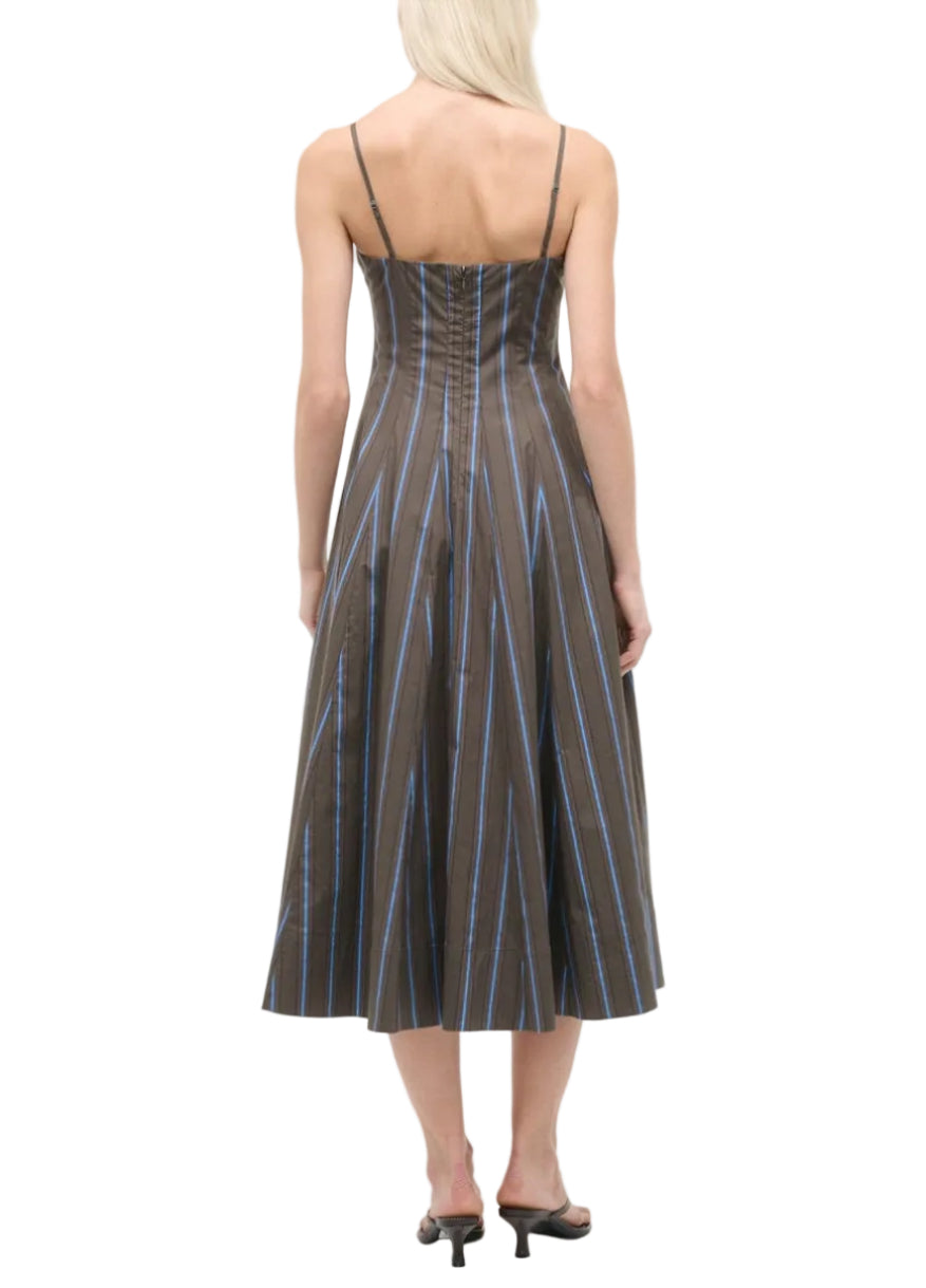 A woman with light hair is seen from the back wearing the Staud Wylie Dress by Staud—a sleeveless, corset-style brown midi dress with blue vertical stripes and spaghetti straps—paired with black heeled sandals.