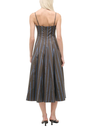 A woman with light hair is seen from the back wearing the Staud Wylie Dress by Staud—a sleeveless, corset-style brown midi dress with blue vertical stripes and spaghetti straps—paired with black heeled sandals.