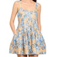 A woman stands with her hands in the pockets of the Cara Cara Ria Dress, a sleeveless beige dress featuring blue floral prints and a square neckline.