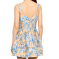 A woman is shown from the back wearing the Cara Cara Ria Dress, a sleeveless beige dress with blue floral prints, a fitted bodice, and pintucked detailing.