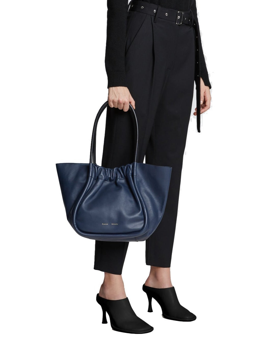 Woman holding a Proenza Schouler Large Ruched Tote bag and wearing a black belt, pants, and heeled shoes, cropped at the waist.