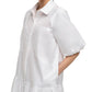 A woman wears the Simkhai Crissy Dress, a loose-fitting white mini dress with a shirting style, puffed sleeves, front and side pockets, and her hands in the side pockets.