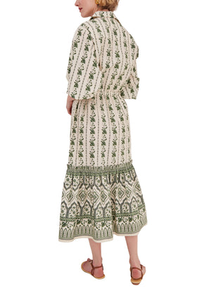 A woman faces away from the camera, wearing the Cara Cara Hutton Dress—a patterned, long-sleeve, ankle-length cream and green dress with a cinched waist—and sandals.