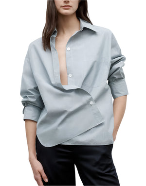 A person wears the TWP New Earl Shirt—an oversized, relaxed, light blue button down in Italian cotton with an asymmetrical closure—paired with black pants and standing with one hand in their pocket.