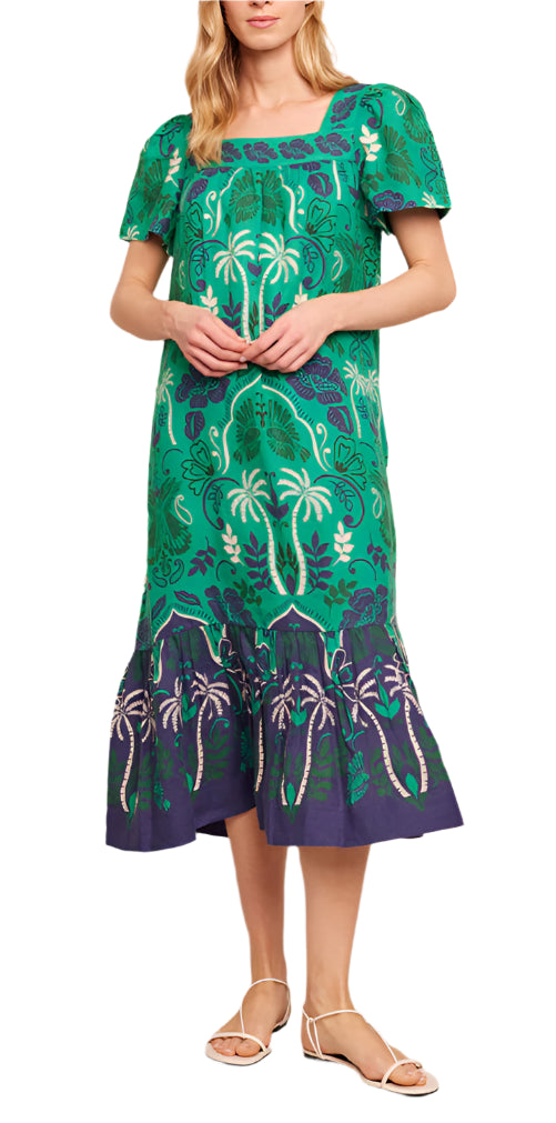 A woman in a Cara Cara Chani Dress, showcasing an island print with green and blue florals and palm tree patterns, stands in sandals with her hands on her midsection.