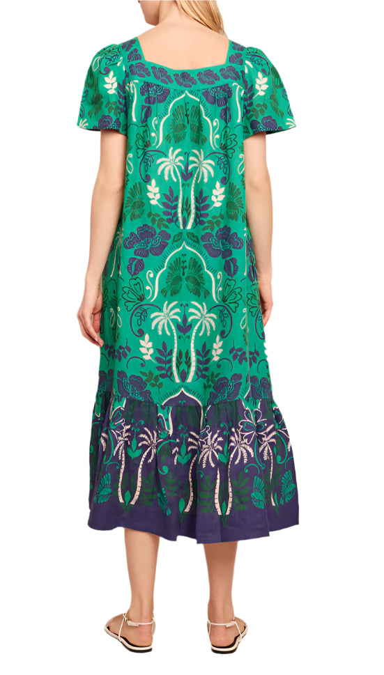 The woman is wearing a Cara Cara Chani Dress by Cara Cara, showcasing an island print with a bohemian vibe. The green and blue patterns create a tropical design that complements the short sleeves beautifully as she is viewed from the back, highlighting the dress's vibrant allure.