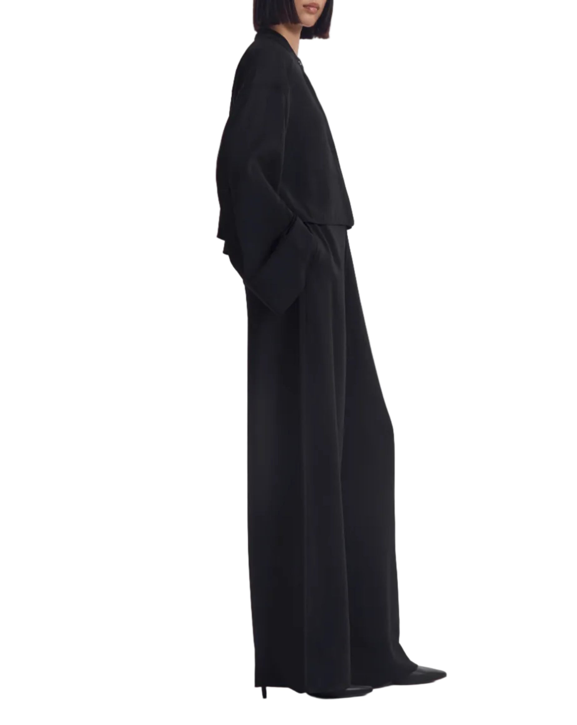 A woman, standing in profile against a white background, wears CO's Wide Leg Crepe Pant as part of a loose-fitting black ensemble with long sleeves.