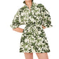 A person wears the Henry Dress by Cara Cara, a button down mini dress with a green leaf and botanical print, rolled sleeves, collared neckline, and cinched waist.
